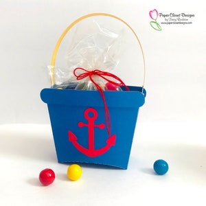 May include: A blue paper party favour box with a red anchor design and a gold handle. The box is filled with colourful sweets and tied with a red ribbon.