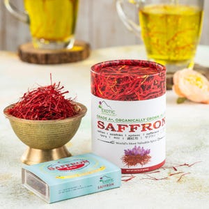 May include: An image showcasing saffron products. A small brass bowl holds a pile of red saffron threads. A cylindrical container and a small blue box, both labeled "SAFFRON", are also present. Two mugs of saffron tea are in the background.