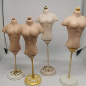 May include: Four miniature dress forms in varying shades of beige and cream. Each form is mounted on a wooden dowel and a decorative base. The bases are white, gold, and cream-colored, with ornate designs. The forms are labeled A, B, C, and D.