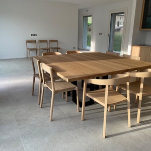 Puede incluir: Una gran mesa de madera con un acabado marrón claro y una base de metal negro. La mesa está rodeada por ocho sillas de madera con un acabado marrón claro. Las sillas están dispuestas en círculo alrededor de la mesa. La mesa y las sillas están en una habitación con paredes blancas y un suelo de baldosas.