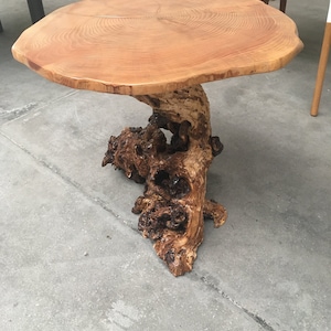 May include: A round wooden table with a natural edge and a unique tree trunk base. The tabletop is light brown with visible wood grain. The base is a dark brown, intricately shaped tree trunk. The table is a rustic, natural design.