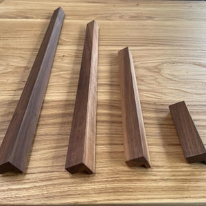 May include: Four wooden furniture legs of different sizes, displayed on a light brown wooden surface. The legs have a geometric, angled design and are a mix of dark and light brown tones. These legs are likely for a table or other furniture.