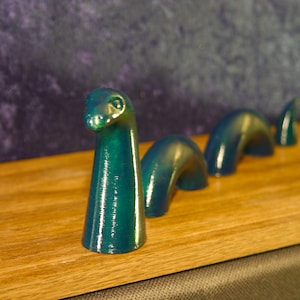 May include: A teal-colored sculpture of the Loch Ness Monster, with a long neck and arched body segments, is displayed on a wooden surface. The sculpture has a glossy finish and is set against a dark, textured background.