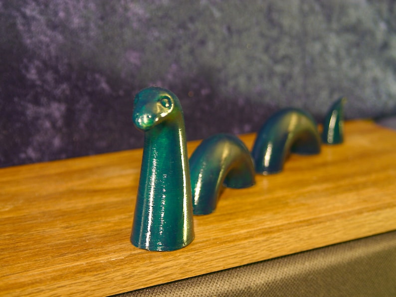 May include: A teal-colored sculpture of the Loch Ness Monster, with a long neck and arched body segments, is displayed on a wooden surface. The sculpture has a glossy finish and is set against a dark, textured background.