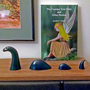 May include: A book titled "The Catalpa Tree Fairy and Other Stories" by Jennifer P. Tanaba, featuring an illustration of a fairy with yellow hair and wings, sitting on a rock in a green forest.  The book is displayed on a wooden shelf with four small, dark green ceramic sculptures.