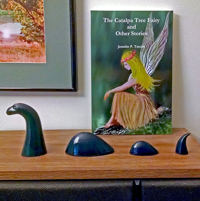 May include: A book titled "The Catalpa Tree Fairy and Other Stories" by Jennifer P. Tanaba, featuring an illustration of a fairy with yellow hair and wings, sitting on a rock in a green forest.  The book is displayed on a wooden shelf with four small, dark green ceramic sculptures.