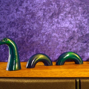 May include: A green, stylized sculpture of the Loch Ness Monster, with its head and humps emerging from the water. The sculpture is set against a purple backdrop and rests on a wooden surface.