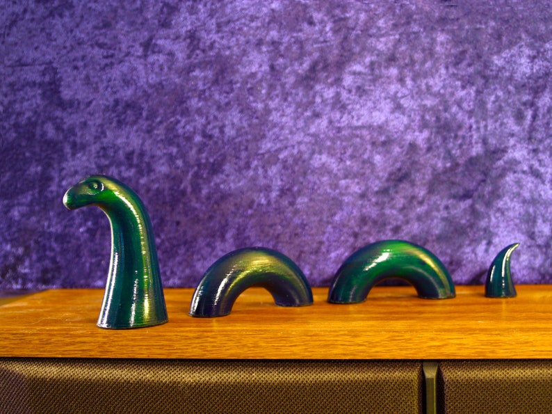 May include: A green, stylized sculpture of the Loch Ness Monster, with its head and humps emerging from the water. The sculpture is set against a purple backdrop and rests on a wooden surface.