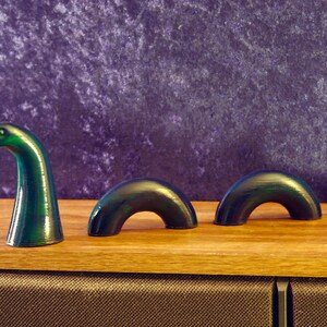 May include: A green and black sculpture of the Loch Ness Monster, with a head and three humps, is displayed on a wooden surface. The sculpture has a glossy finish and is set against a purple background. The artwork is a whimsical representation of the legendary creature.