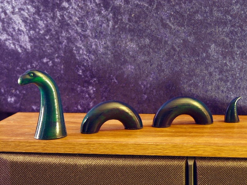 May include: A green and black sculpture of the Loch Ness Monster, with a head and three humps, is displayed on a wooden surface. The sculpture has a glossy finish and is set against a purple background. The artwork is a whimsical representation of the legendary creature.