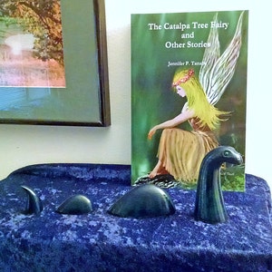May include: A book titled "The Catalpa Tree Fairy and Other Stories" by Jennifer P. Tanab, featuring a fairy with wings and a flower crown, sitting on a log in a green forest. Three dark green ceramic sculptures of a mythical creature are in the foreground.