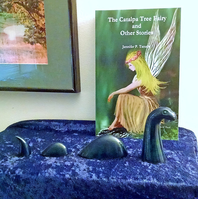 May include: A book titled "The Catalpa Tree Fairy and Other Stories" by Jennifer P. Tanab, featuring a fairy with wings and a flower crown, sitting on a log in a green forest. Three dark green ceramic sculptures of a mythical creature are in the foreground.