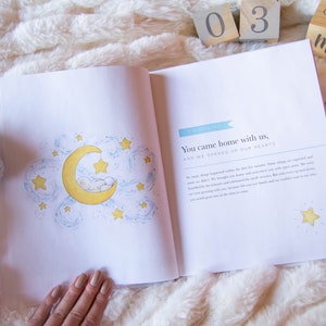 May include: An open baby book with a blue and white illustration of a crescent moon with stars and a sleeping baby. The text on the page reads "You came home with us, and we opened up our hearts."  The book is surrounded by a white fluffy blanket, wooden blocks that read "03" and "Weeks/Month", and two crocheted elephant booties.