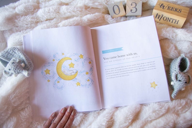 May include: An open baby book with a blue and white illustration of a crescent moon with stars and a sleeping baby. The text on the page reads "You came home with us, and we opened up our hearts."  The book is surrounded by a white fluffy blanket, wooden blocks that read "03" and "Weeks/Month", and two crocheted elephant booties.