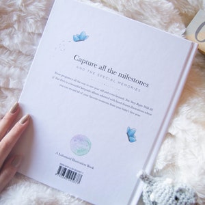 May include: A white book with the text "Capture all the milestones and the special memories" on the cover. The book is lying on a white fluffy surface with wooden blocks that say "03 weeks" and "month".