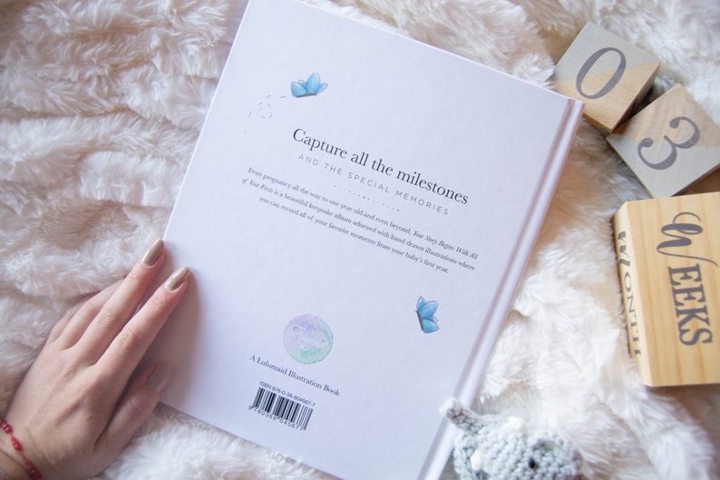 May include: A white book with the text "Capture all the milestones and the special memories" on the cover. The book is lying on a white fluffy surface with wooden blocks that say "03 weeks" and "month".