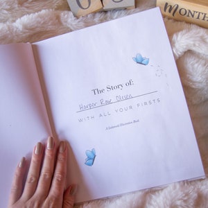 May include: An open book with a white cover and the title "The Story of: Harper Rose Olsen with all your firsts" written in black ink. The book is on a white fluffy surface with two small gray crocheted elephant toys.