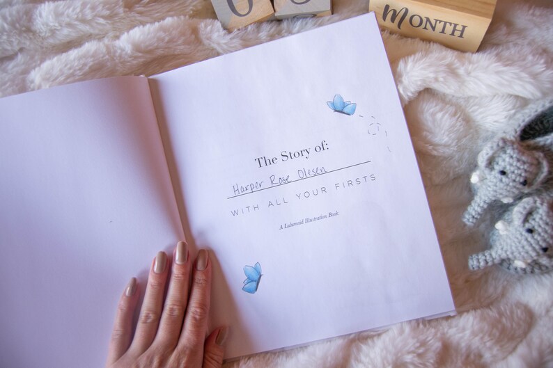 May include: An open book with a white cover and the title "The Story of: Harper Rose Olsen with all your firsts" written in black ink. The book is on a white fluffy surface with two small gray crocheted elephant toys.