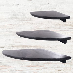 May include: Three black wooden shelves in a half-circle shape, mounted on a white wall. The shelves are made of dark wood and have a rustic finish.