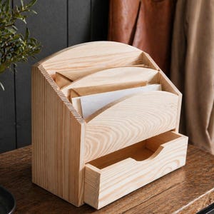 Può includere: Un organizer da scrivania in legno chiaro con un cassetto. L'organizer ha tre fessure curve per contenere carte e buste. Il cassetto è aperto, rivelando l'interno. La venatura del legno è visibile.