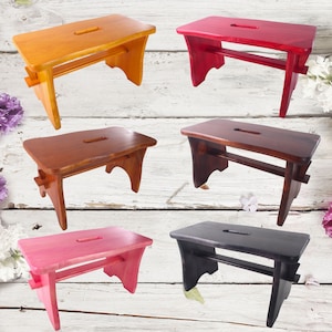 Op de afbeelding: Vijf houten krukjes in verschillende kleuren, waaronder naturel hout, rood, bruin, roze en zwart. Elk krukje heeft een eenvoudig ontwerp met een rechthoekige zitting en twee poten aan elke kant.