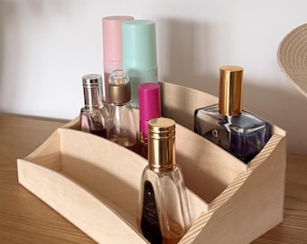 May include: A wooden tiered organizer holding various perfume bottles and cosmetic items. The organizer is made of light-colored wood and has three sections. The bottles are in various shapes and sizes, with gold and silver caps, and pink, blue, and clear containers.