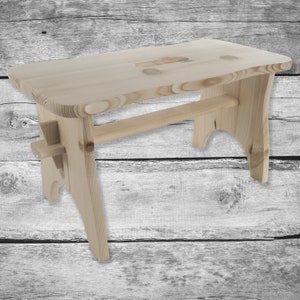 May include: A natural wood step stool with a simple design. The stool has a rectangular top with a small cutout and two legs that are slightly curved.