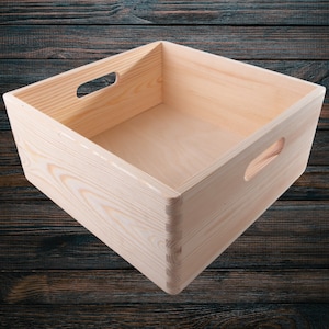 Puede incluir: Una caja de madera simple con dos asas en la parte superior y una en el lateral. La caja está vacía y hecha de madera clara.