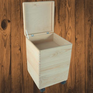 Könnte beinhalten: Eine hellfarbene Holzaufbewahrungsbox mit einem Scharnierdeckel, der geöffnet ist, um das Innere freizulegen. Die Box ist rechteckig, auf kleinen Rädern montiert und scheint aus unbehandeltem Holz zu bestehen.