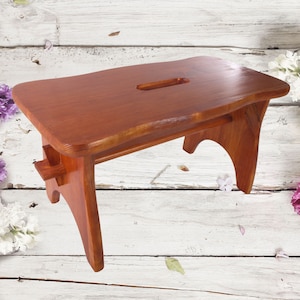 May include: A solid wood step stool with a natural wood finish. The stool has a rectangular top with a cutout handle and two curved legs.