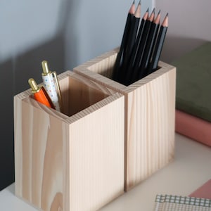 Peut inclure: Deux porte-crayons en bois, l'un avec des crayons noirs et l'autre avec des stylos colorés. Les porte-crayons sont en bois clair et ont une finition naturelle.