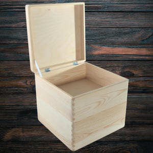 Peut inclure: Une boîte en bois clair inachevée avec un couvercle à charnière, légèrement entrouvert. La boîte est carrée et présente un grain de bois visible. L'intérieur est vide. Le fond est un motif de grain de bois foncé. Idéal pour le rangement.