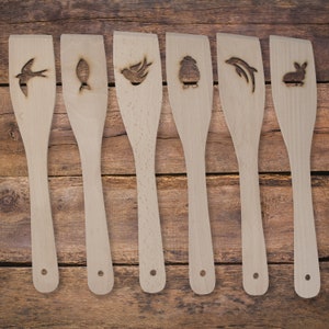 May include: Seven wooden kitchen spatulas with different designs carved into them. The designs include a bird, a fish, a rabbit, a dolphin, a bird, and a flower.