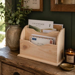 May include: A natural wood letter rack holding envelopes and a magazine. It's on a wooden surface, next to a vase with foliage, a candle, and a small brass dish. A framed sign above reads "IT'S GOOD TO BE HOME."