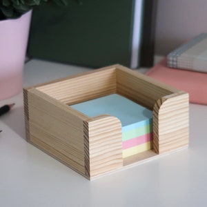 Puede incluir: Un organizador de escritorio de madera con una pila de notas adhesivas de colores pastel. El organizador está hecho de madera clara y tiene una forma cuadrada.