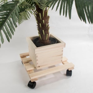 May include: A wooden plant stand with wheels, featuring a square planter box. The stand is made of natural wood and has a light brown finish. The planter box is empty and ready to hold a plant.