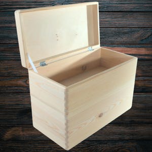 Può includere: Una scatola di legno color chiaro con coperchio incernierato, parzialmente aperto. La scatola è rettangolare con finitura a venatura naturale. Il coperchio è fissato con cerniere metalliche e un supporto bianco. La scatola è vuota.