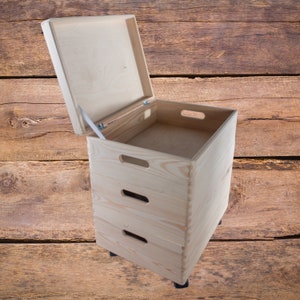 Può includere: Una scatola di legno per riporre oggetti con un coperchio incernierato e tre cassetti. La scatola è realizzata in legno naturale e ha una finitura marrone chiaro. La scatola è su ruote e ha un aspetto rustico.