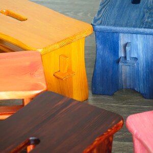 May include: Five wooden stools in different colors: orange, yellow, brown, blue, and pink. The stools are all made of wood and have a simple design.
