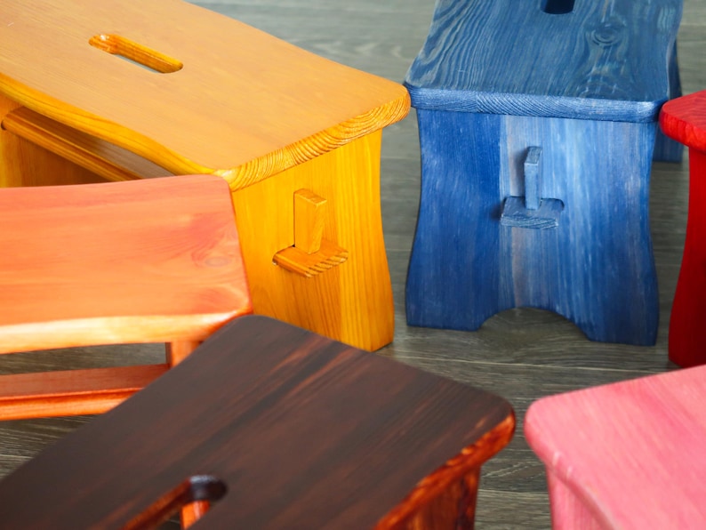May include: Five wooden stools in different colors: orange, yellow, brown, blue, and pink. The stools are all made of wood and have a simple design.