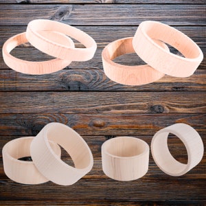 Pode incluir: Seis anéis de madeira de tamanhos diferentes, empilhados e dispostos sobre uma superfície de madeira castanho-escura. Os anéis são todos de madeira clara e têm um acabamento liso e polido.