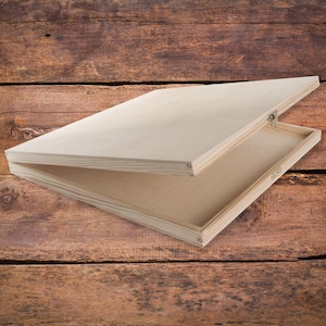 Pode incluir: Uma caixa de madeira com tampa articulada, aberta para revelar o interior vazio. A caixa é feita de madeira clara e tem um acabamento natural.