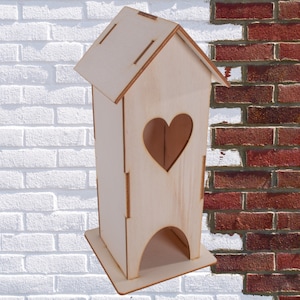 May include: A wooden house-shaped box with a heart-shaped cutout on the front. The box is light brown and has a small opening at the bottom.