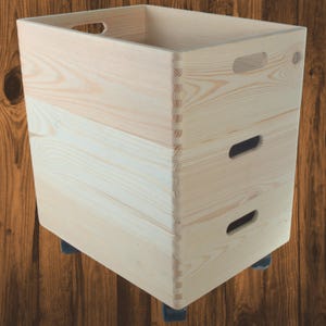 May include: A light-colored wooden storage bin with a rectangular shape. The bin has finger-jointed corners, cut-out handles, and two smaller cut-out handles on the side. It sits on four small black wheels.