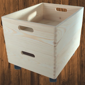 Op de afbeelding: Een lichtgekleurde houten opbergkrat met uitgesneden handgrepen aan de zijkanten. De krat is rechthoekig en staat op vier kleine zwarte wielen. De houtnerf is zichtbaar.