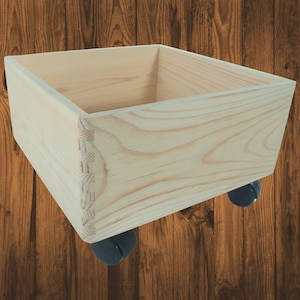 Puede incluir: Una caja de madera de color claro con forma cuadrada, con veta de madera visible y uniones de cola de milano. La caja está montada sobre cuatro pequeñas ruedas negras, diseñadas para facilitar la movilidad. La caja está vacía.