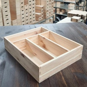 May include: A rectangular light-colored wooden organizer tray featuring four distinct interior compartments of varying sizes. The tray is constructed with visible wood grain patterns and joined corners, providing a structured storage solution for small items.