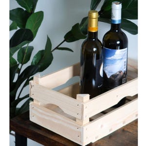 May include: Two wine bottles in a light wooden crate. One bottle has a gold cap and a yellow label, the other has a white and blue label. The crate is on a dark wooden surface, with green leaves in the background.