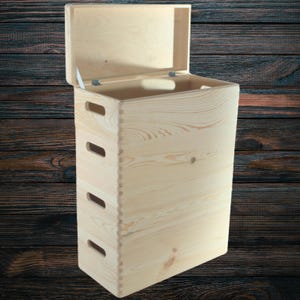 Op de afbeelding: Een lichtgekleurde houten opbergkist met een scharnierend deksel, met vier rechthoekige handgrepen aan de voorkant. De kist is geconstrueerd met zwaluwstaartverbindingen en heeft een natuurlijke houtnerf. Het deksel is gedeeltelijk open.