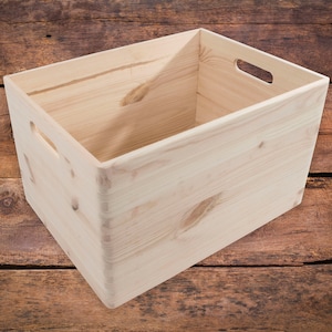 Puede incluir: Una caja de almacenamiento de madera natural con dos asas en los lados. La caja está vacía y hecha de madera sin tratar.